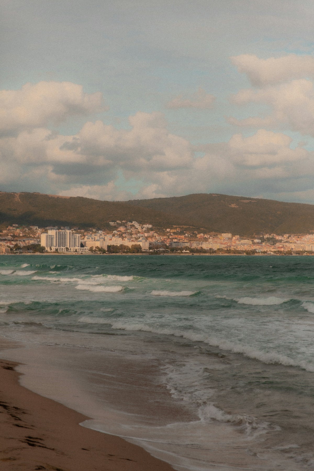 Waves crash on a sandy beach with city in background.